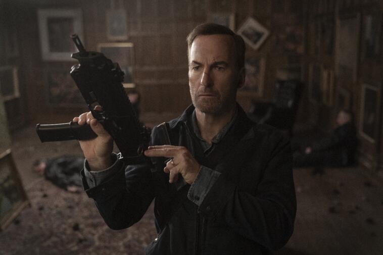 Schauspieler Bob Odenkirk verkörpert Nobody im gleichnamigen Film und hält eine automatische Waffe, einsatzbereit, hoch